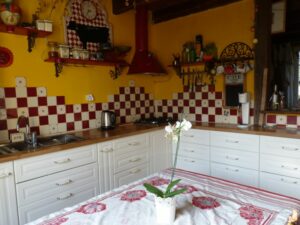 Logement de Melgven ,La cuisine blanche avec le carrelage mural balnc et rouge et la table avec une nappe avec des fleurs rouges et une orchidée posée dessus