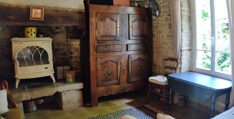 salon avec poele blanc dans la cheminée, fenêtre a droite avec une vielle table grisée en dessous. On voit un meuble breton clouté , une lampe à côté et une chaise avec un coussin